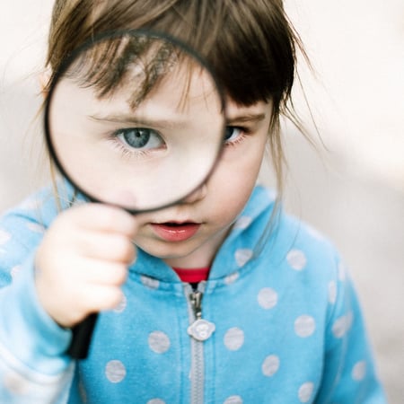 Child in blue sweatshirt with white polkadots peering through a large magnifying glass