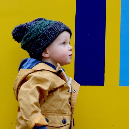 Boy with knit blue hat standing in front of a yellow wall looking off into the distance